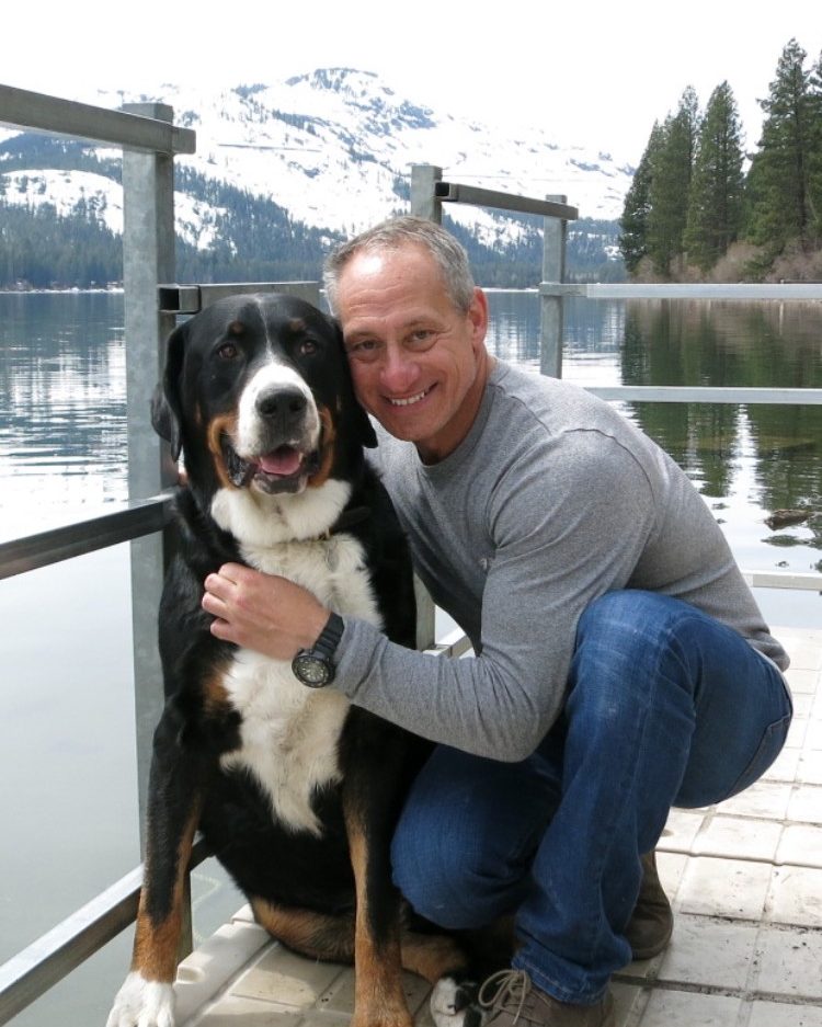 Ben crouching beside Murray, a large Swiss mountain dog, on a lakeside dock with snowy peaks behind