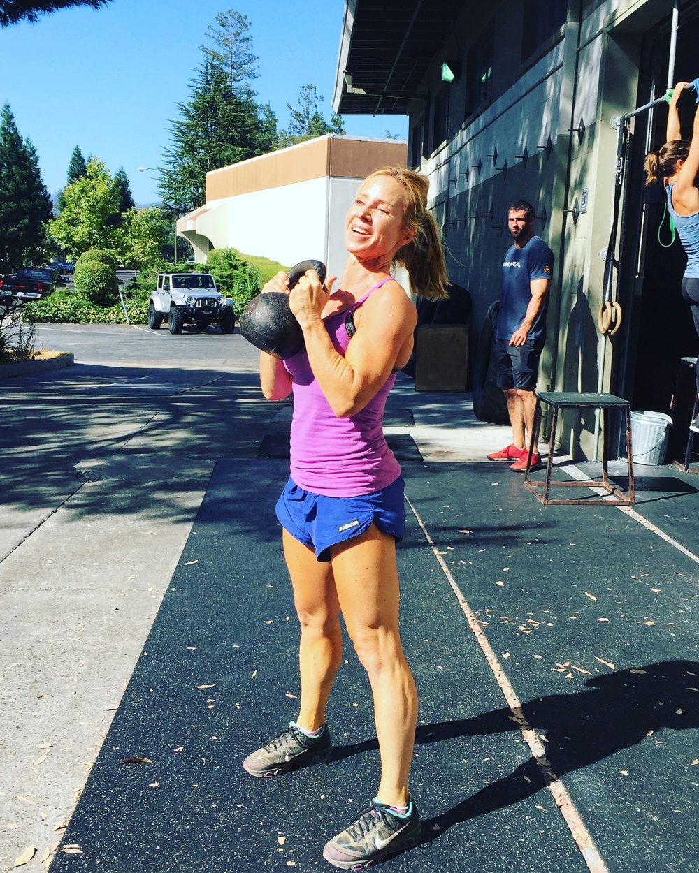 Loree holding a kettlebell at her shoulder outside a gym, smiling