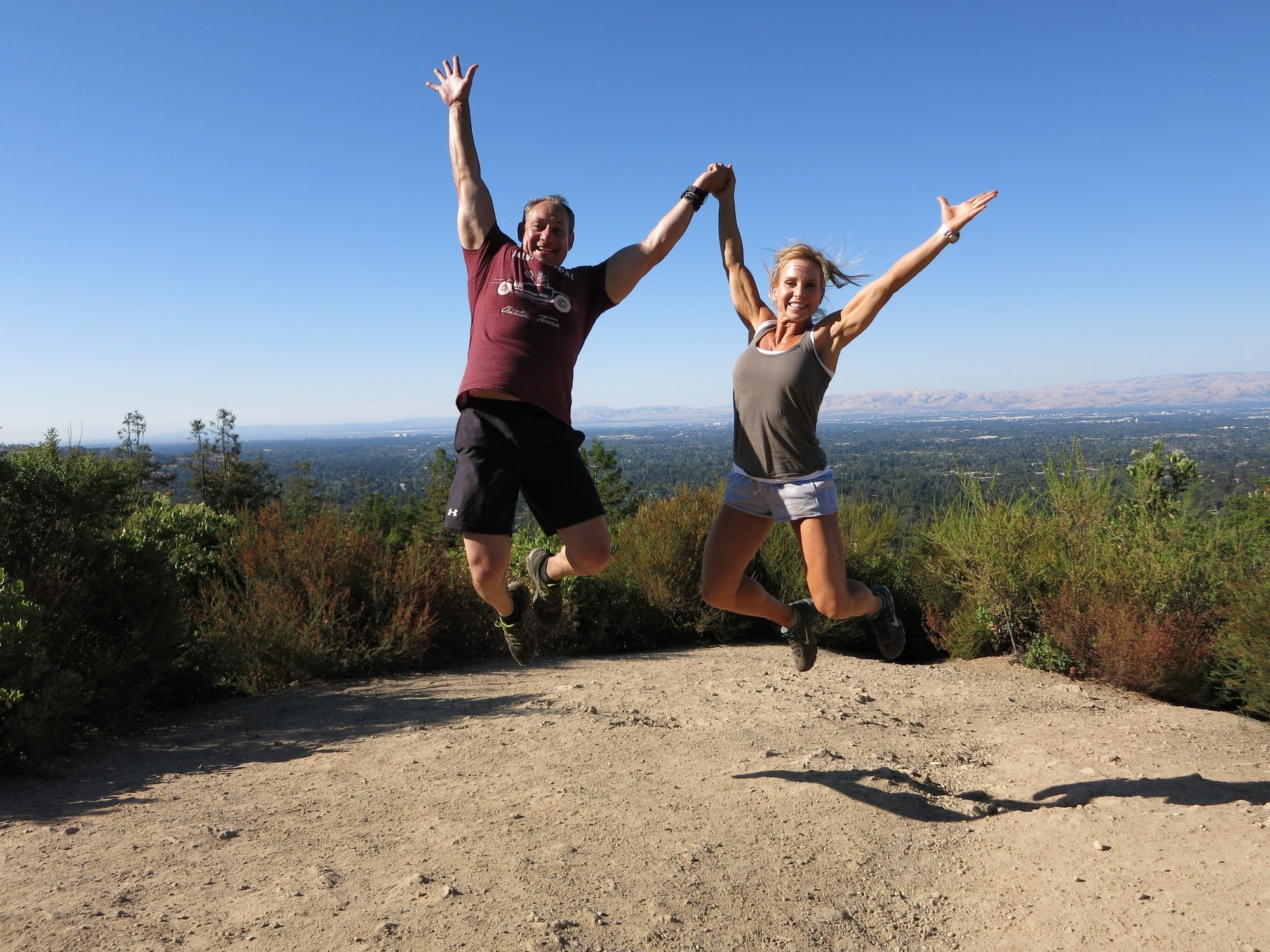 Ben and Loree jumping in the air on a hilltop trail, arms raised, valley far below
